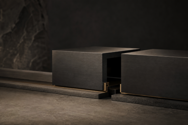 Dark editorial still life of two charcoal metal blocks aligned across a subtle hidden break in a handoff track